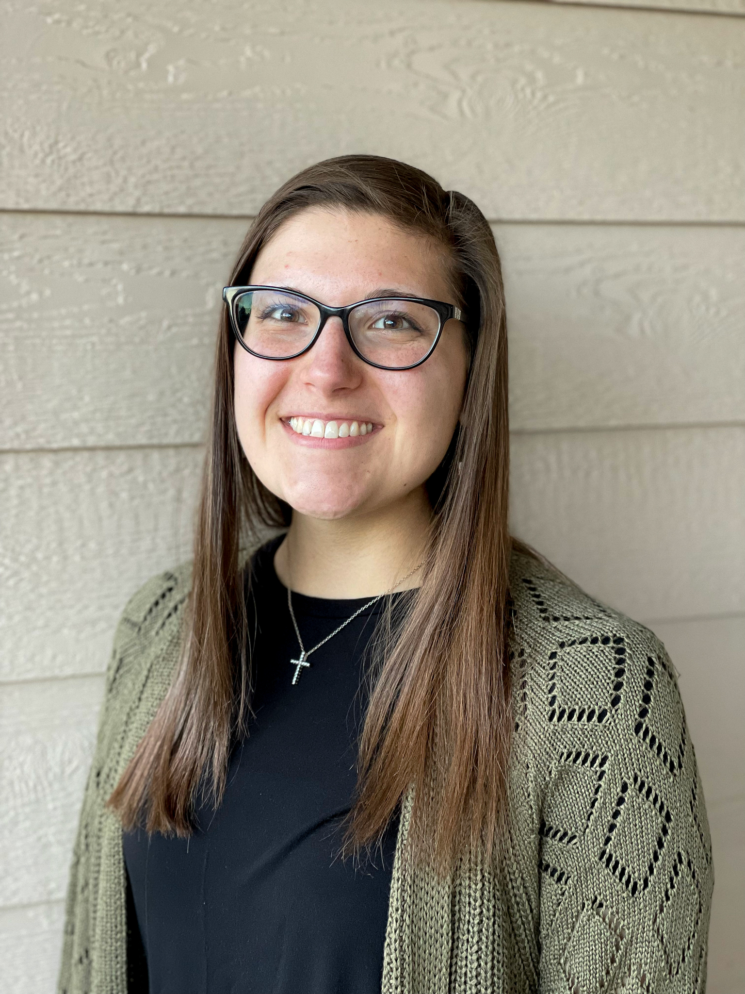 Headshot of Andreanna Haritopoulos, a female young-adult with long brown hair and glasses wearing a green cardigan with a black shirt and cross necklace